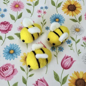 Puede incluir: Tres adornos de abeja de fieltro amarillo con rayas negras y alas blancas están dispuestos sobre una tela estampada con flores. La tela presenta rosas rosas, margaritas azules, girasoles amarillos y otras flores coloridas.