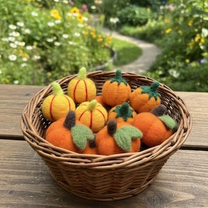 Może przedstawiać: Wiklinowy kosz wypełniony ręcznie robionymi filcowymi dyniami w różnych odcieniach pomarańczowego i żółtego. Dynie mają zielone łodygi i liście. Kosz stoi na drewnianej powierzchni z rozmytym ogrodem w tle.