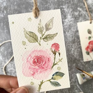 May include: A watercolor painting of a pink rose with green leaves and a bud on a white background. The painting is on a small card with a hole punched in the top and a twine string attached.