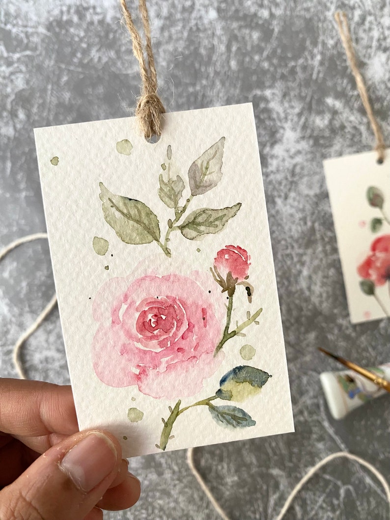 May include: A watercolor painting of a pink rose with green leaves and a bud on a white background. The painting is on a small card with a hole punched in the top and a twine string attached.