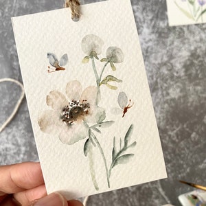 May include: A watercolor painting of a white flower with green leaves and a butterfly. The painting is on a white card with a hole punched in the top and a string attached.