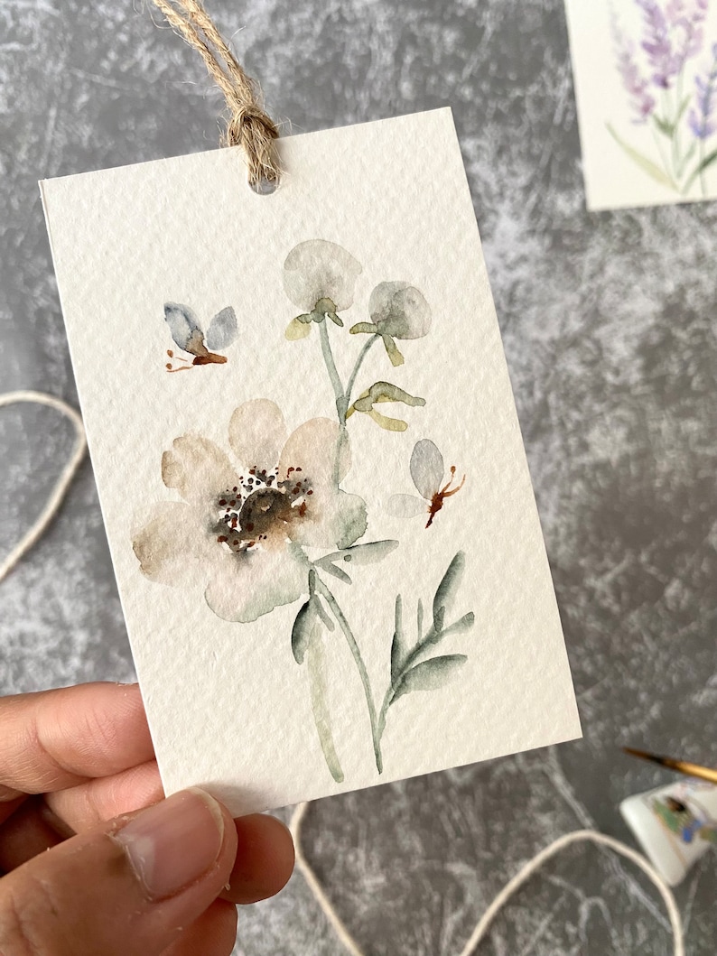 May include: A watercolor painting of a white flower with green leaves and a butterfly. The painting is on a white card with a hole punched in the top and a string attached.