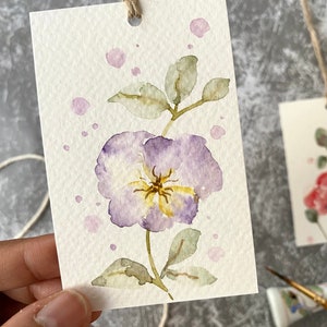 May include: A watercolor painting of a purple flower with yellow center and green leaves on a white background with pink watercolor splatters.