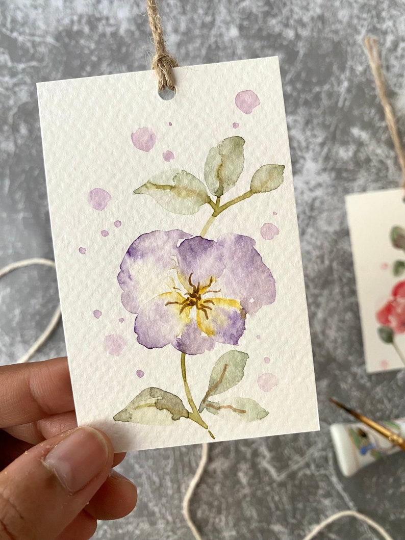 May include: A watercolor painting of a purple flower with yellow center and green leaves on a white background with pink watercolor splatters.