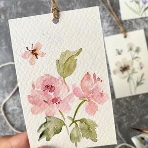 May include: A watercolor painting on a white card with a pink flower, green leaves, and a butterfly. The card has a hole punched in the top and a string attached.