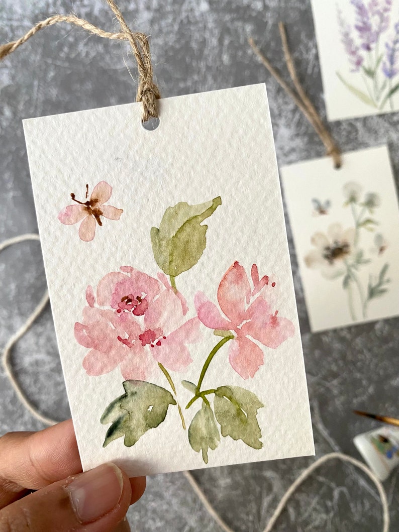 May include: A watercolor painting on a white card with a pink flower, green leaves, and a butterfly. The card has a hole punched in the top and a string attached.
