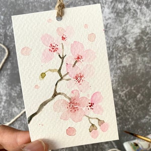 May include: A watercolor painting of a delicate pink cherry blossom branch with a few scattered pink petals on a white background. The painting is on a small card with a hole punched in the top and a twine string attached.