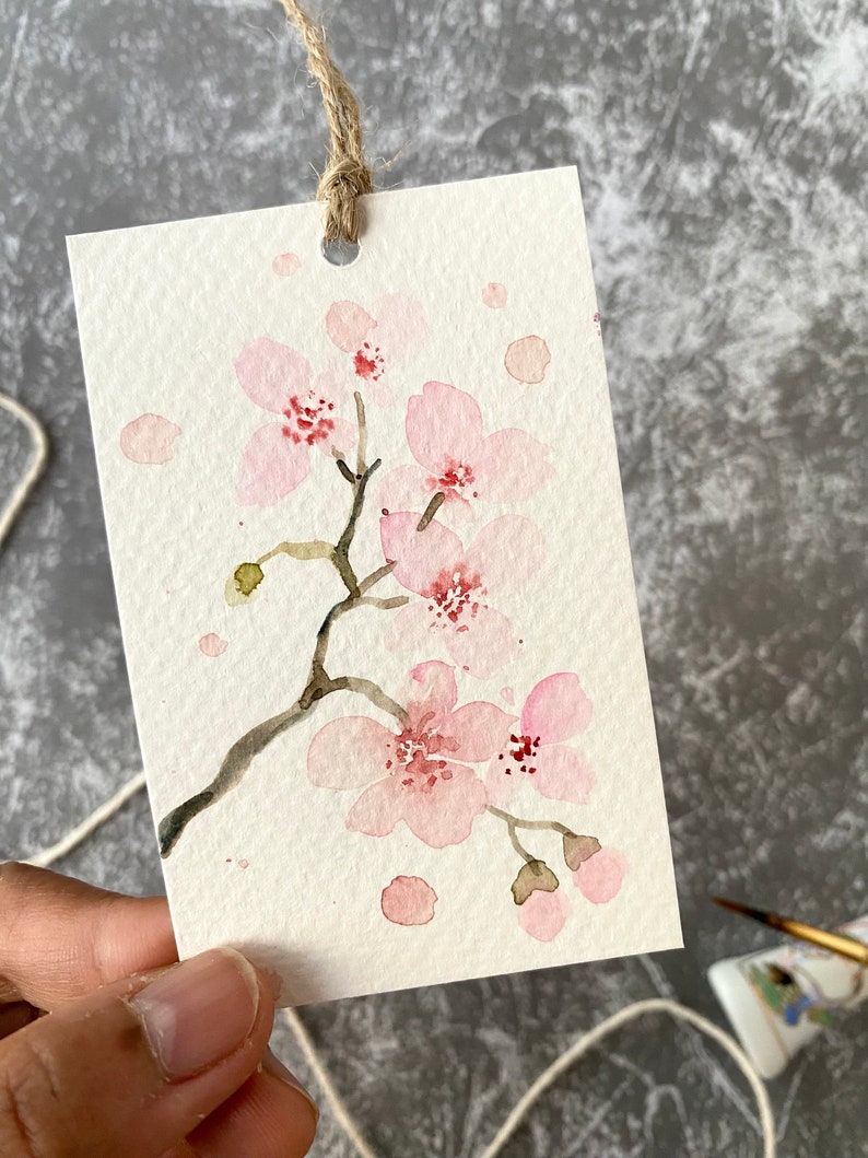May include: A watercolor painting of a delicate pink cherry blossom branch with a few scattered pink petals on a white background. The painting is on a small card with a hole punched in the top and a twine string attached.