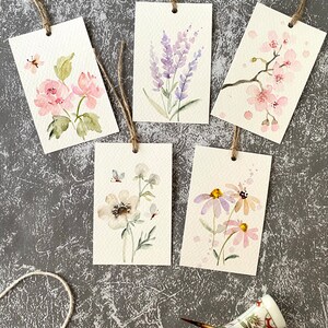 May include: Five watercolor painted gift tags with floral designs. The tags are tied with twine and feature different flowers, including lavender, cherry blossoms, and daisies.
