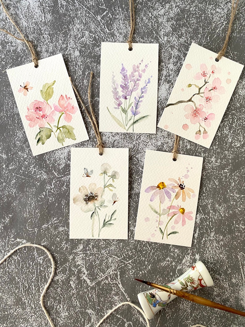 May include: Five watercolor painted gift tags with floral designs. The tags are tied with twine and feature different flowers, including lavender, cherry blossoms, and daisies.