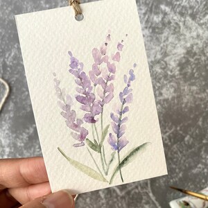 May include: A watercolor painting of lavender flowers on a white card with a hole punched in the top. The card is tied with twine.