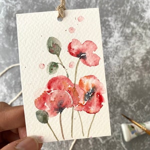 May include: Watercolor painting of red poppies on a white card with a brown string attached. The painting is a simple, delicate depiction of the flowers.