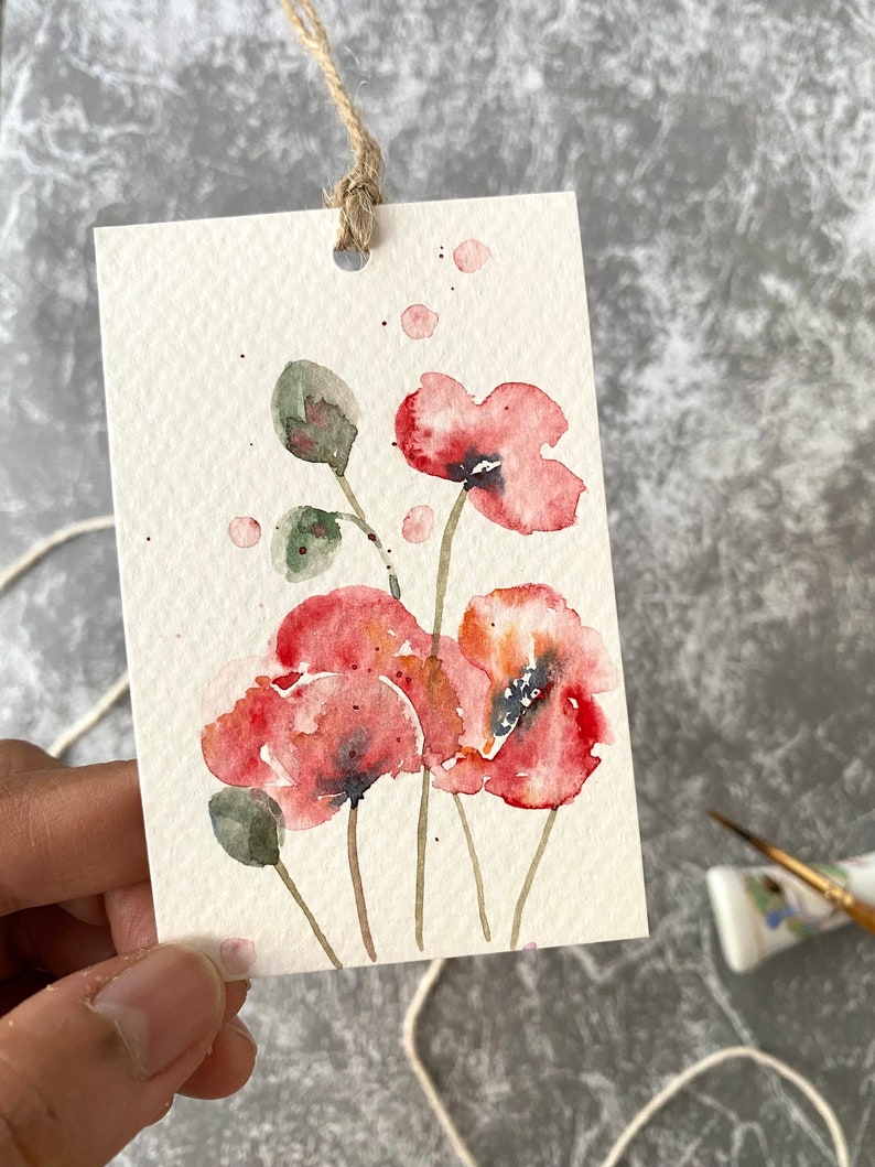 May include: Watercolor painting of red poppies on a white card with a brown string attached. The painting is a simple, delicate depiction of the flowers.