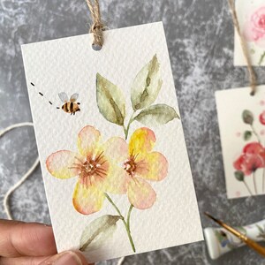May include: A watercolor painting of two yellow flowers with pink accents on a white background. A small bee is flying above the flowers. The painting is on a white card with a hole punched in the top and a string attached.