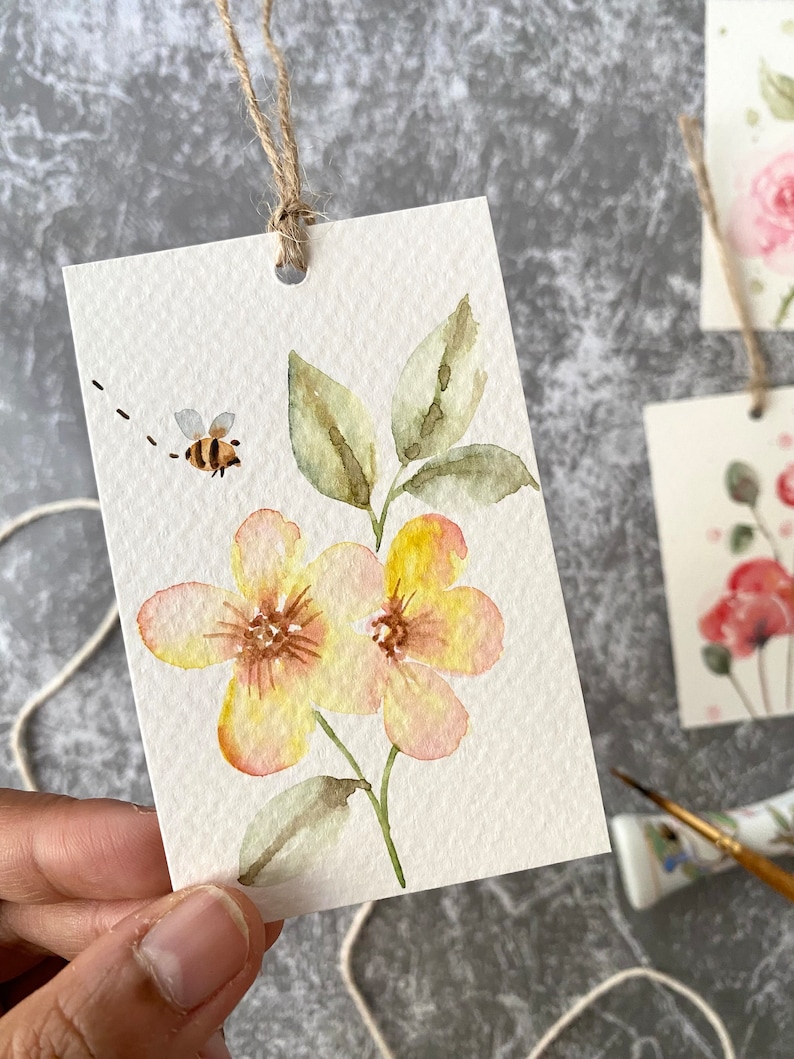 May include: A watercolor painting of two yellow flowers with pink accents on a white background. A small bee is flying above the flowers. The painting is on a white card with a hole punched in the top and a string attached.