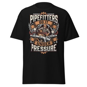 Puede incluir: Una camiseta negra de manga corta con una ilustración gráfica de un instalador de tuberías trabajando en tubos de metal con una llave inglesa. El diseño incluye el texto "PIPEFITTERS DO IT UNDER PRESSURE" rodeado de engranajes, manómetros y alas.