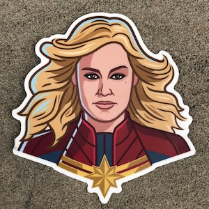 Può includere: Un'illustrazione a fumetti di una supereroina che indossa un costume rosso, blu e oro con una stella sul petto. Ha i capelli biondi e un'espressione seria sul viso.