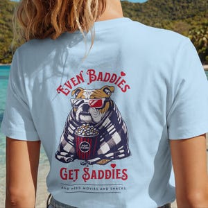 Może przedstawiać: Jasnoniebieski t-shirt z grafiką buldoga w okularach 3D, owiniętego w koc w kratę i trzymającego wiaderko popcornu. Napis głosi "EVEN BADDIES GET SADDIES" oraz "NEED MOVIES AND SNACKS" wykonany czerwoną czcionką.