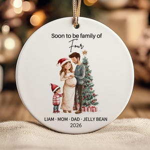 Può includere: Un ornamento rotondo in ceramica bianca con un cordino dorato e l'illustrazione di una famiglia vicino a un albero di Natale. Il testo recita Soon to be family of Four, LIAM, MOM, DAD, JELLY BEAN e 2026. La famiglia include un bambino.