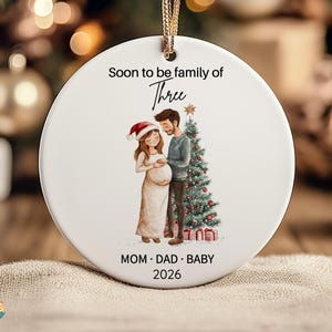 Puede incluir: Un adorno navideño redondo de cerámica blanca que cuelga de un cordón dorado. Presenta una ilustración de una persona embarazada y su pareja junto a un árbol de Navidad decorado. El texto dice: Soon to be family of Three, MOM · DAD · BABY, 2026.