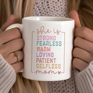 以下が含まれることがあります： ピンクのハートが付いたカラフルな枠の中に「she is strong fearless warm loving patient selfless mom」という文字が書かれた白いセラミック製のコーヒーカップです。