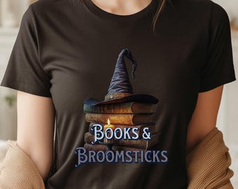 May include: A black t-shirt featuring a graphic of a stack of old books topped with a dark blue pointed hat. A lit candle sits in front of the books. The text "BOOKS & BROOMSTICKS" is printed in a stylized blue font below the candle and books.