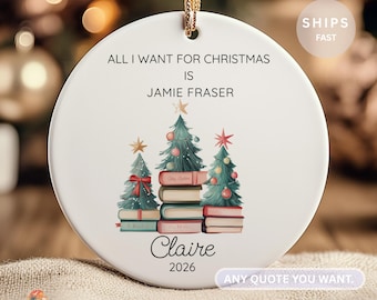 Puede incluir: Adorno navideño de cerámica blanca con cordón dorado. El adorno tiene el texto «ALL I WANT FOR CHRISTMAS IS JAMIE FRASER» y «Claire 2026». Presenta ilustraciones de árboles de Navidad sobre pilas de libros.
