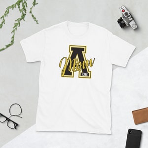 May include: White t-shirt with a black letter A with a gold outline. The letter A has the word "Mom" written in a gold cursive font inside of it. The number "06" is written in black below the letter A.
