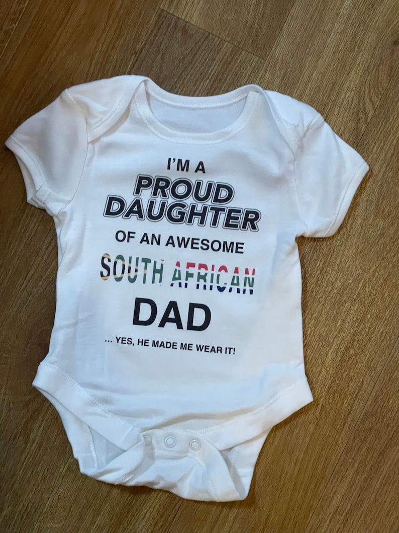 May include: White baby onesie with short sleeves. The onesie has the text "I'M A PROUD DAUGHTER OF AN AWESOME SOUTH AFRICAN DAD ... YES, HE MADE ME WEAR IT!" printed on the front in black and colorful lettering. The onesie has snap closures at the bottom.