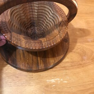 Puede incluir: Un cuenco de fruta de madera con un dise&ntilde;o tallado y un asa. El cuenco est&aacute; hecho de madera marr&oacute;n oscura y tiene una forma redonda.