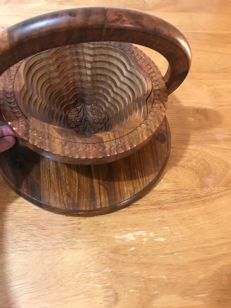 Puede incluir: Un cuenco de fruta de madera con un dise&ntilde;o tallado y un asa. El cuenco est&aacute; hecho de madera marr&oacute;n oscura y tiene una forma redonda.