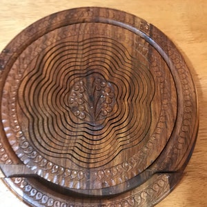 Puede incluir: Un cesto de fruta redondo de madera extensible con un dise&ntilde;o floral tallado en el centro. El cesto est&aacute; hecho de madera marr&oacute;n oscura y tiene un acabado liso y pulido.