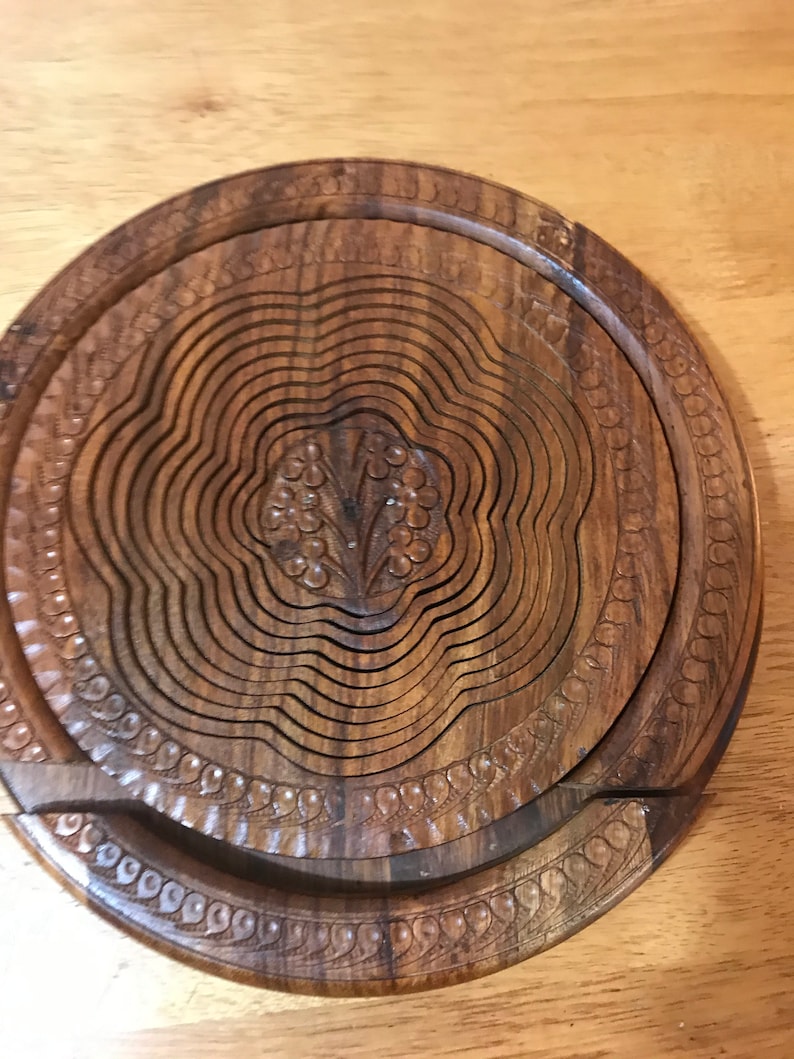 Puede incluir: Un cesto de fruta redondo de madera extensible con un dise&ntilde;o floral tallado en el centro. El cesto est&aacute; hecho de madera marr&oacute;n oscura y tiene un acabado liso y pulido.