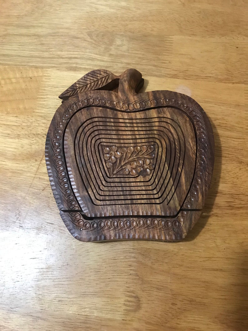 May include: A wooden apple-shaped fruit bowl with intricate carvings. The bowl is made of dark brown wood and has a leaf design on the top. The bowl is designed to be collapsible, with multiple layers that can be stacked together.