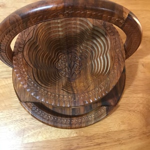 Puede incluir: Un frutero de madera con un dise&ntilde;o tallado y un asa. El frutero est&aacute; hecho de m&uacute;ltiples capas de madera que se pueden plegar para su almacenamiento.