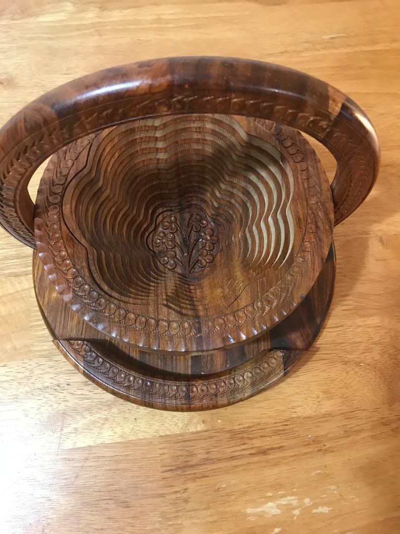 Puede incluir: Un frutero de madera con un dise&ntilde;o tallado y un asa. El frutero est&aacute; hecho de m&uacute;ltiples capas de madera que se pueden plegar para su almacenamiento.