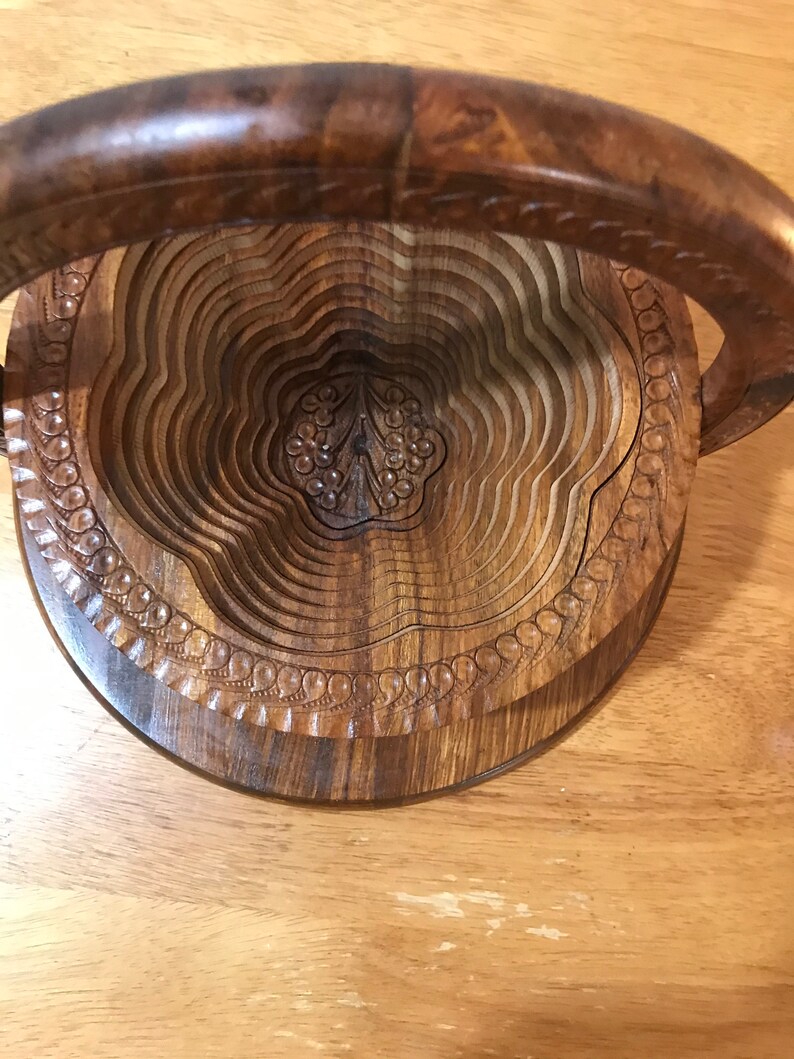 Puede incluir: Un frutero de madera con un dise&ntilde;o tallado y un asa. El frutero es de madera oscura y tiene forma circular.