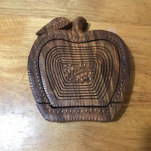 May include: A wooden apple-shaped fruit bowl with intricate carvings. The bowl is made of dark brown wood and has a leaf design on the top. The bowl is designed to be collapsible, with multiple layers that can be stacked together.
