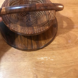 Puede incluir: Un cuenco de fruta de madera con asa. El cuenco est&aacute; hecho de m&uacute;ltiples capas de madera y tiene un dise&ntilde;o tallado. El cuenco est&aacute; sentado sobre una base de madera.