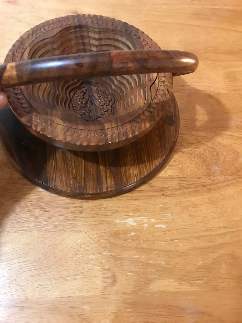 Puede incluir: Un cuenco de fruta de madera con asa. El cuenco est&aacute; hecho de m&uacute;ltiples capas de madera y tiene un dise&ntilde;o tallado. El cuenco est&aacute; sentado sobre una base de madera.