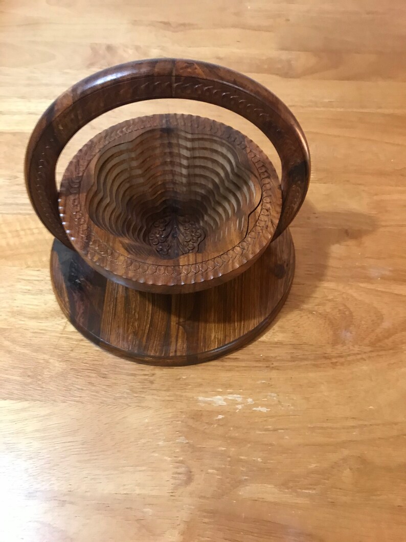 Puede incluir: Un frutero de madera con un dise&ntilde;o tallado y un asa. El frutero est&aacute; hecho de madera oscura y tiene un dise&ntilde;o en capas.