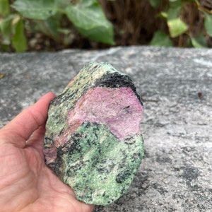 May include: A rough-cut gemstone with a mix of colors. The stone features a prominent section of vibrant pink, contrasted by areas of deep black and speckled green. The overall texture is rugged and natural, showcasing the raw beauty of the mineral.