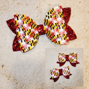 May include: Three red, white, and black bow hair clips with a Maryland flag pattern. The largest bow is in the center and the two smaller bows are below it.