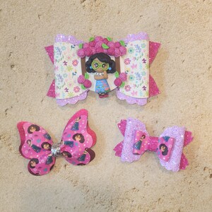May include: Three pink and purple hair bows with a floral design and a cartoon girl wearing glasses. The bows are decorated with glitter and have a variety of patterns.