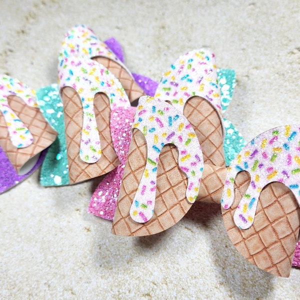 May include: Three ice cream cone hair bows with white, pink, purple and teal glitter. The bows are made of felt and have a waffle cone texture. The ice cream is white with colourful sprinkles.