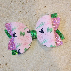 May include: A pink and green hair bow with a witch theme. The bow has a pink and white patterned fabric with the words "wicked" and a witch's hat. The bow is decorated with green glitter.