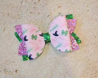 May include: A pink and green hair bow with a witch theme. The bow has a pink and white patterned fabric with the words "wicked" and a witch's hat. The bow is decorated with green glitter.