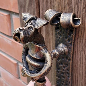 Könnte beinhalten: Bronzefarbener Türgriff mit Drachendesign. Der Griff zeigt eine detaillierte Drachenfigur mit offenem Mund und gewundenem Körper. Der Griff ist auf einer dunklen, strukturierten Metallplatte und einer Holztür montiert. Der Hintergrund umfasst eine Ziegelmauer.