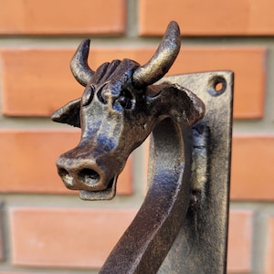 May include: A decorative, metal cow head door knocker. The cow head is bronze-colored with gold accents on the horns and ears. The knocker is mounted on a rectangular plate with two holes for mounting. The background is a red brick wall.
