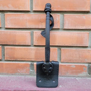 May include: A black, hand-forged metal fire poker with a decorative handle and a shovel-shaped end. The poker has a twisted design element and is set against a brick wall background.